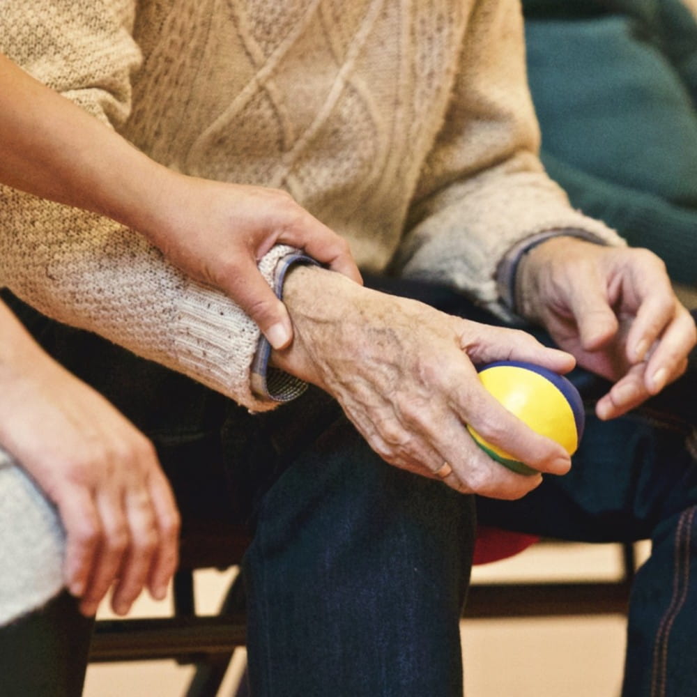 An elderly person, mostly out of shot, holding a small blue and yellow ball in one hand. Another person's hand is resting on their wrist, about to guide them to move the ball.