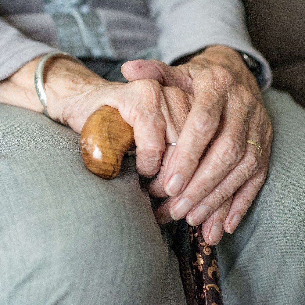 An elderly person's hands folded over each other, one hand holding the handle of a walking stick.