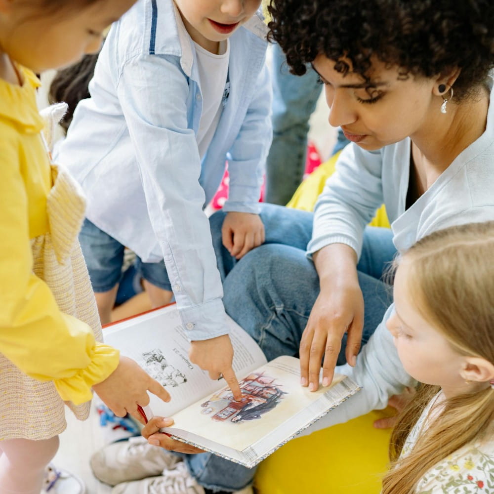 An open book with two children and an adult pointing at a picture. A third child on the right is looking at the picture.