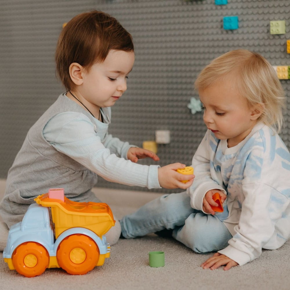 Two toddlers playing on the floor. There is a toy truck in blue and orange in front of them.
