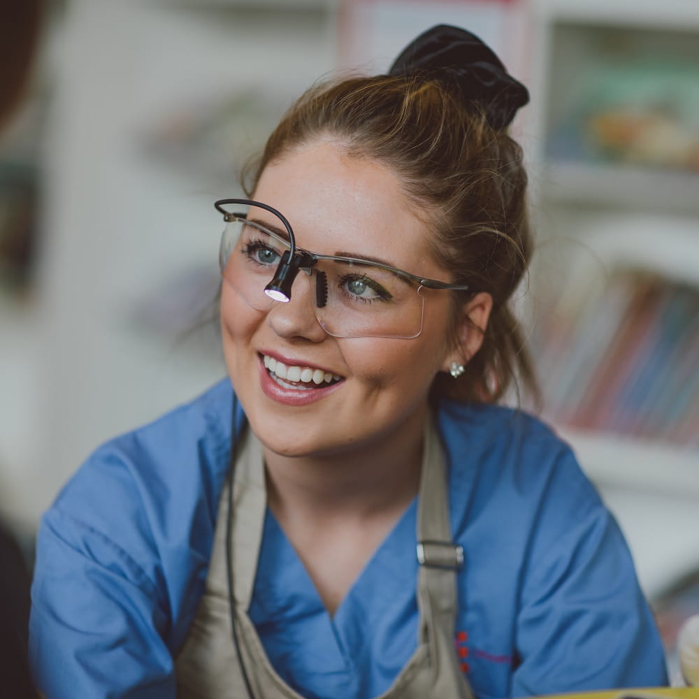A young woman looking to the left of the photo, wearing safety goggles with a torch on the bridge and a blue medical tunic.
