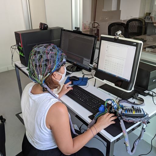 A person wearing an EEG cap is sitting with their back to the photographer. In front of them is a computer screen and keyboard and they are using the computer mouse.