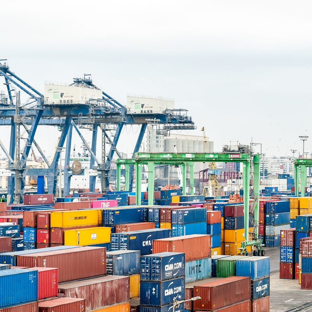 Multicoloured shipping containers sitting on a dock, with a large blue crane in the background.