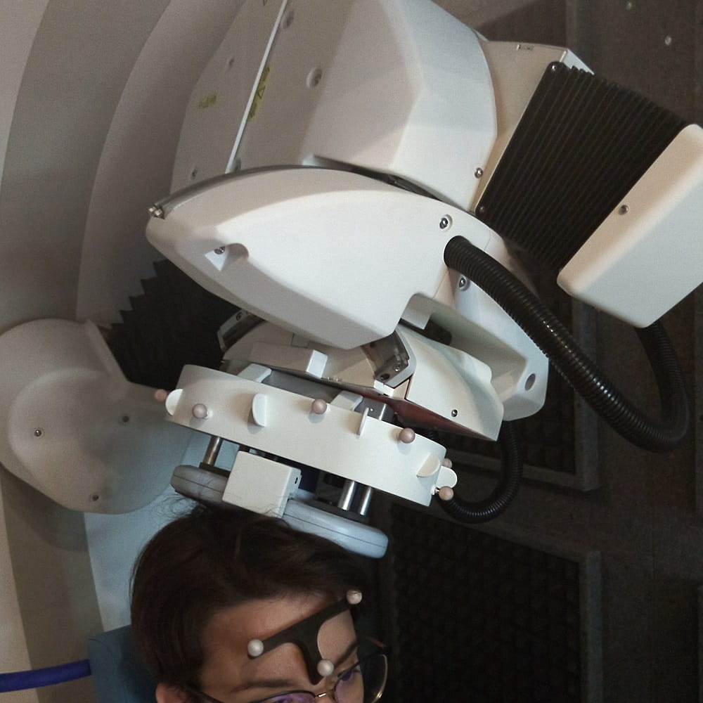In the bottom left of the photo is a person sitting wearing a pair of glasses. Above their head is a large white machine, with a grey ring from it on the person's head.