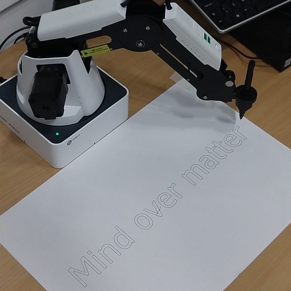 A black and white robot arm holding a pen over a piece of paper that has "Mind Over Matter" written on it.