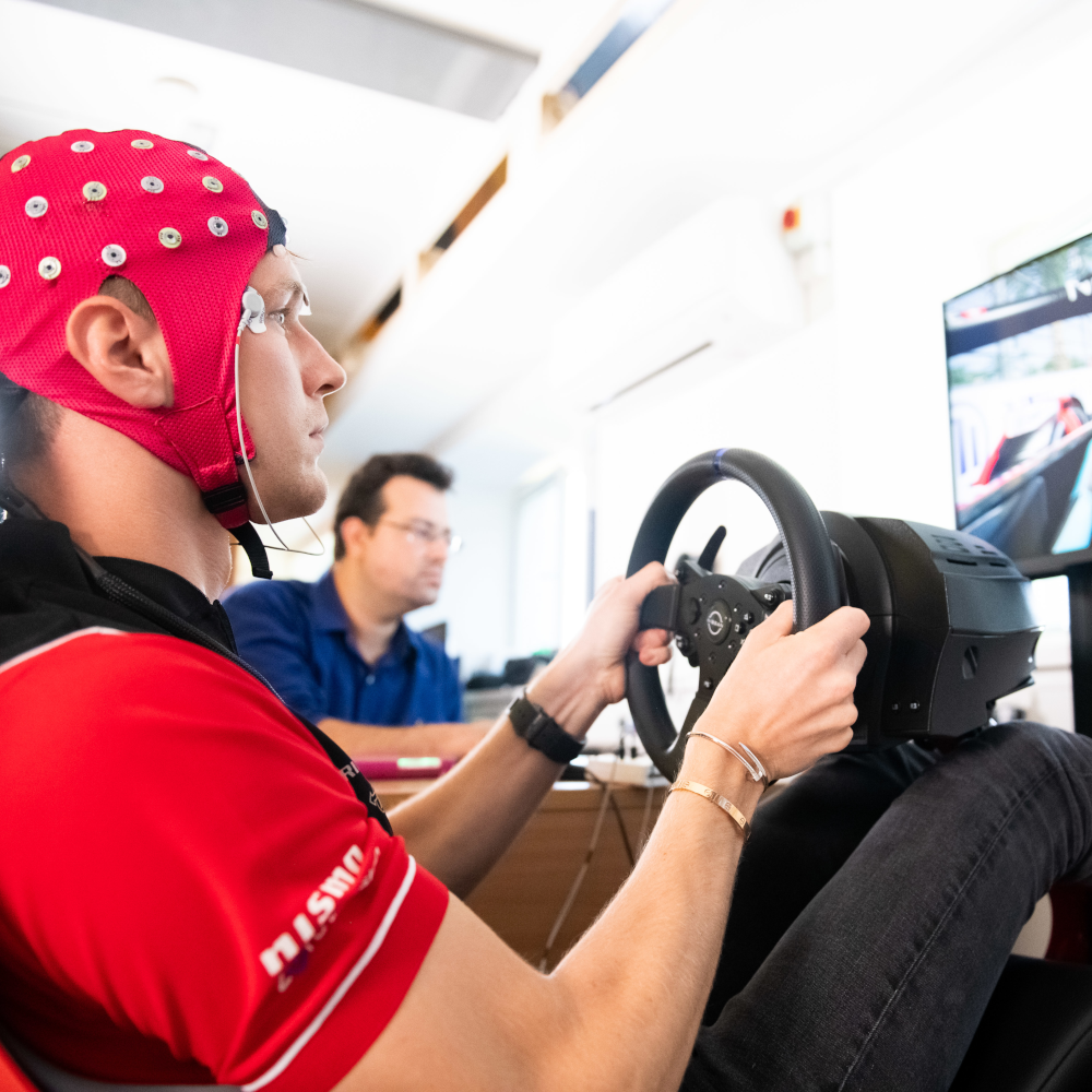 A man wearing a red EEG cap, holding a steering wheel and looking at a screen while a man in a blue shirt sits in the background.