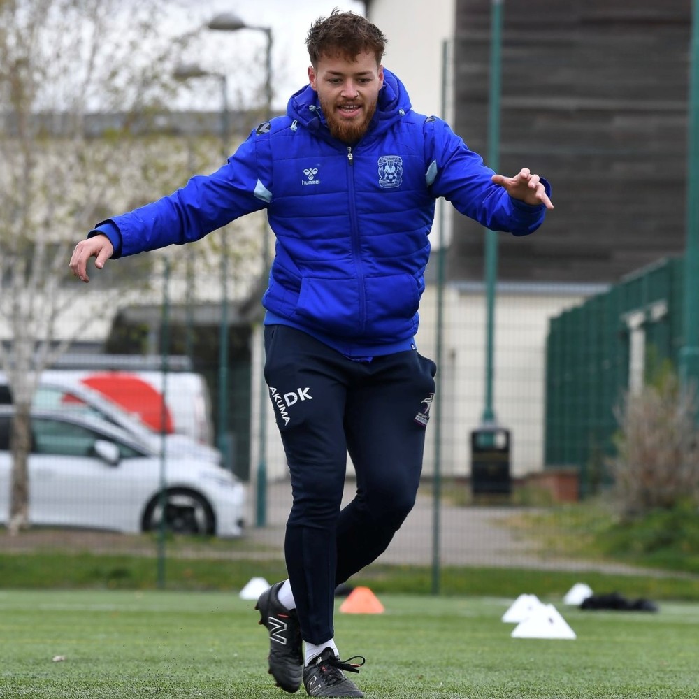 SRES graduate Daniel Kiernan, standing outside on a sports pitch showing an exercise. There are cars, a building and a fence in the background.