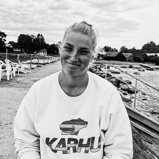 A greyscale photo of Sports Performance and Coaching graduate Helen Hunt, standing on a beach front with sheds and chairs in the background.