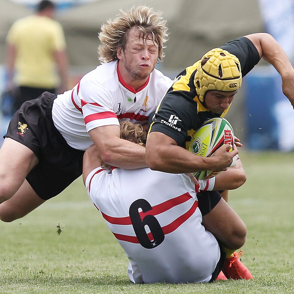 Two rugby players in white shirts with red lines, tackling another player in a black and yellow shirt and a yellow cap on his head, who is holding the rugby ball tucked under his arm.