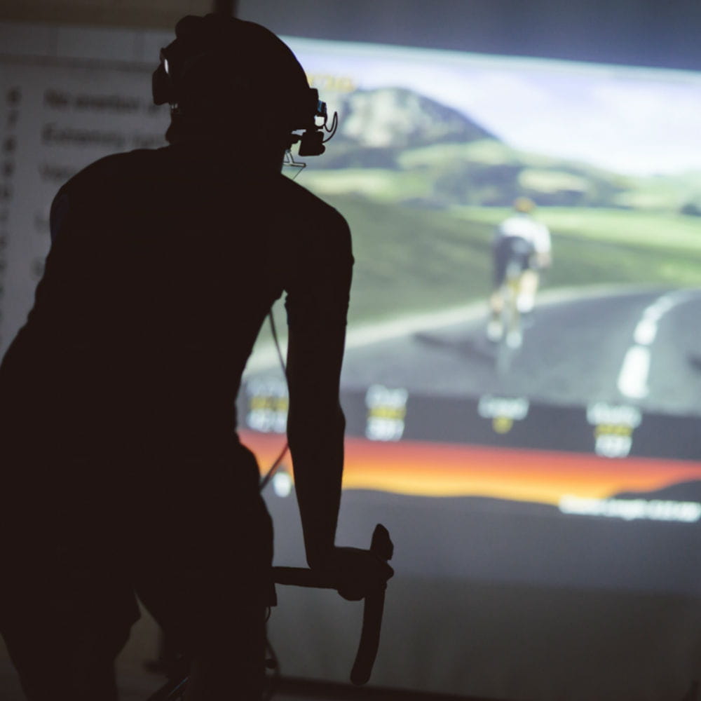 A silhouette of a person on a bike wearing a camera on their head, a video screen in front of them is showing a computer model of a cyclist on a road.