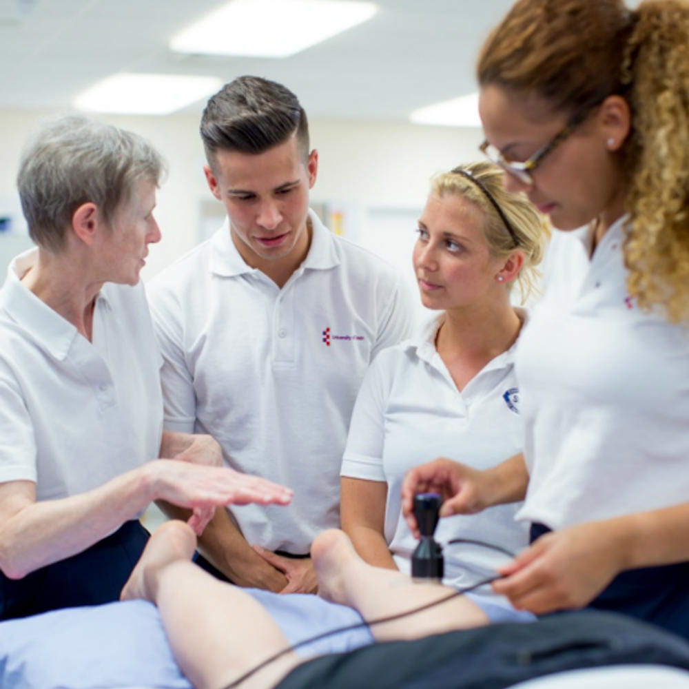 A group of three students and a teacher standing around the end of a bed, with the student on the right holding an ultrasound wand against the leg of a person lying on the bed, mostly out of shot.