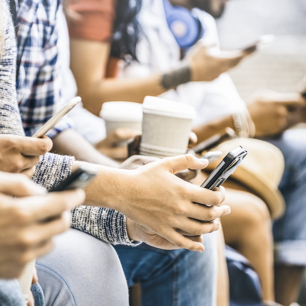 A row of people, their heads out of shot, sitting leaning forward with phones and coffee cups in their hands.