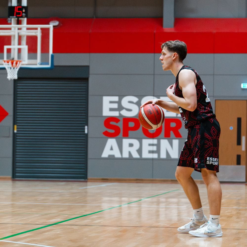 Rebels Mens Basketball Player bouncing basketball on court with text behind 'Essex Sport Arena'