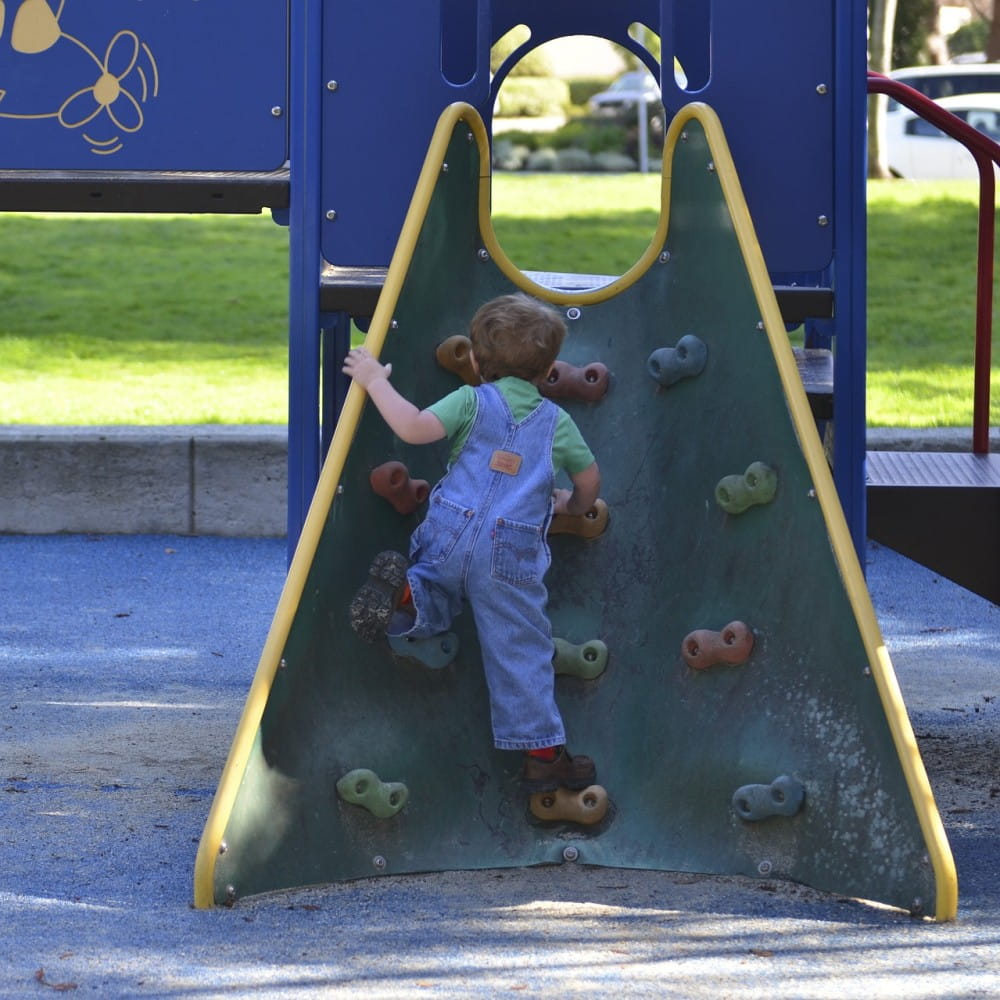 A toddler in dungarees climbing up a children's climbing wall on the side of a play frame.