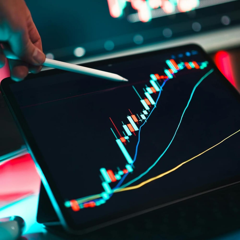 A person out of shot, pointing an iPad pencil at a table. The tablet screen displays a forex chart in light blue and red.