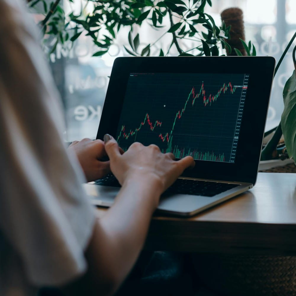 A person, mostly out of shot, sitting at a laptop on a desk. The laptop has a financial chart showing dips and rises.