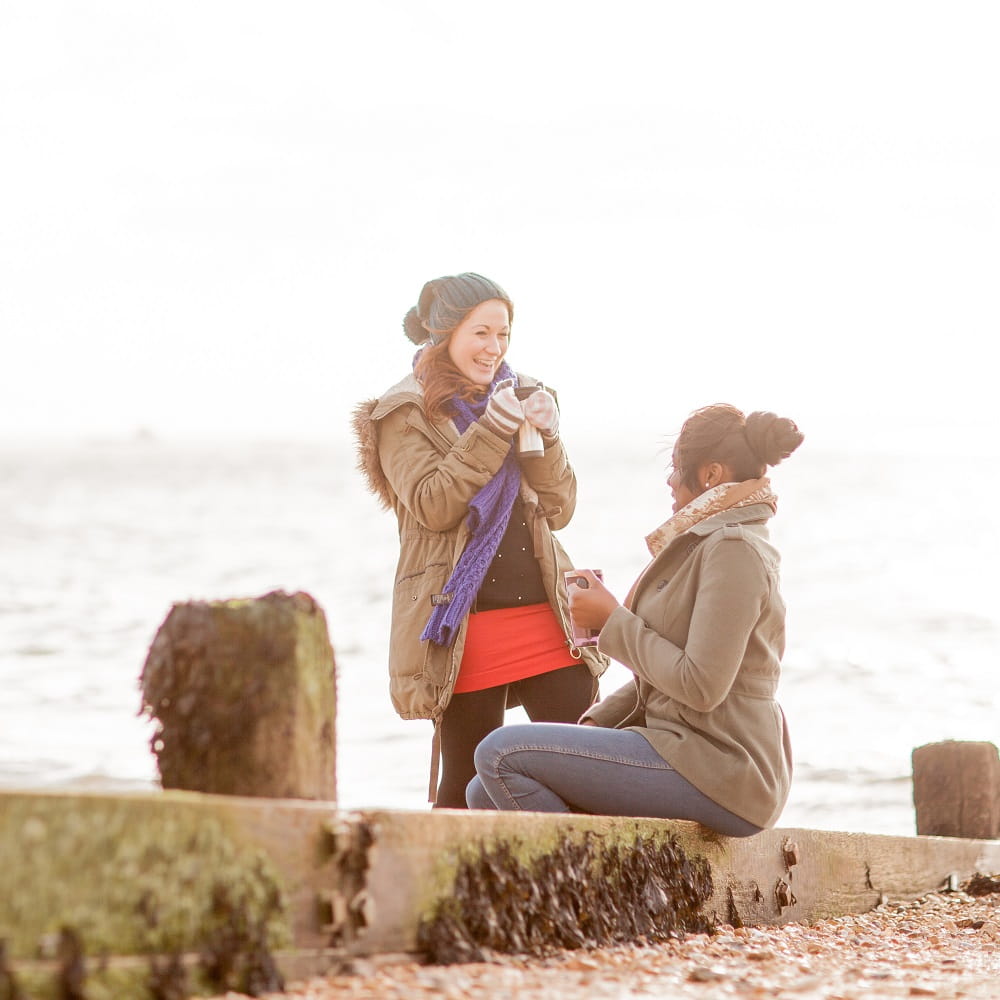 Two women, one standing and one sitting on a wooden groyne, holding travel mugs, with a pale sea and sky in the background.