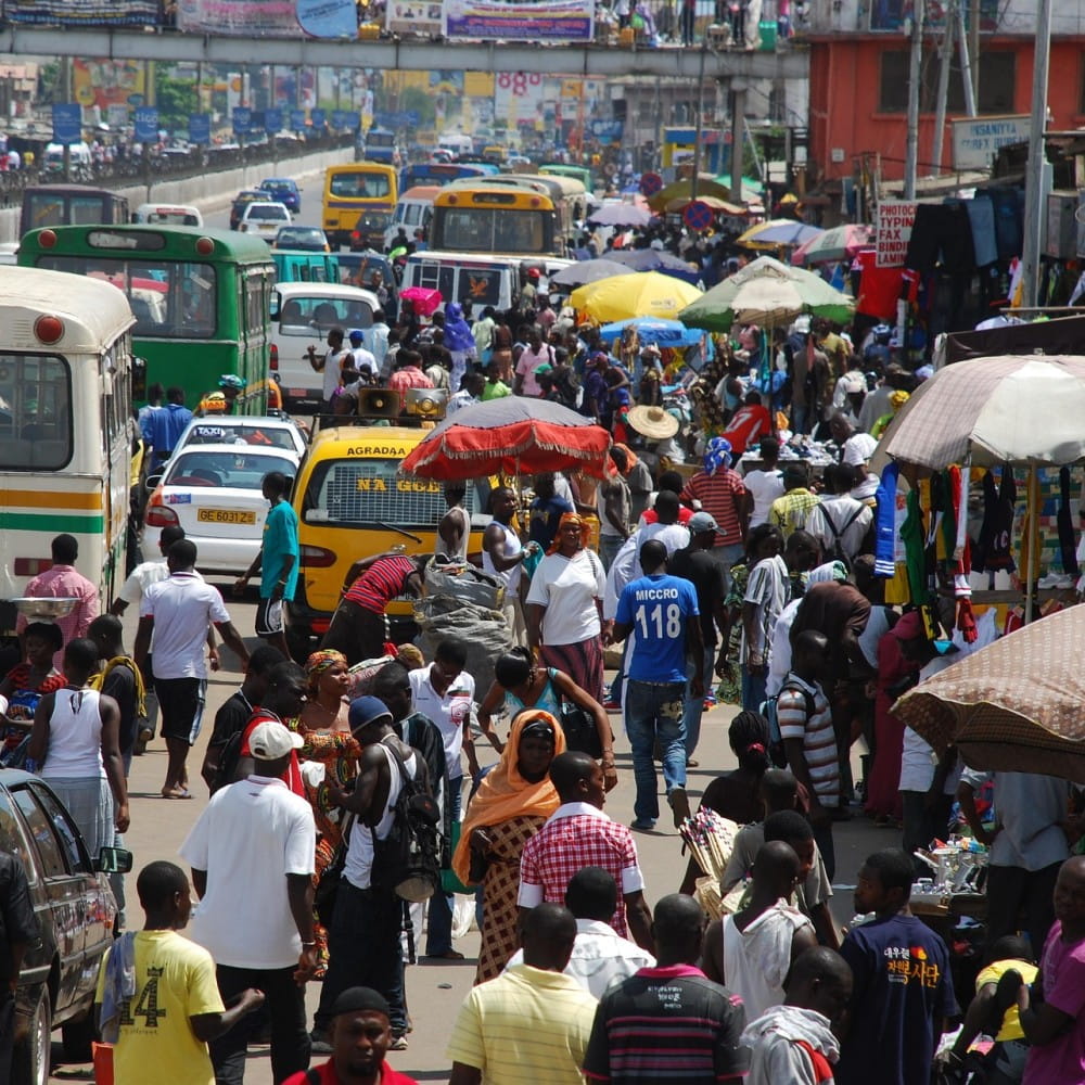 A street in an African city, with cars and buses on the left and people walking on the right, past buildings and market stalls.