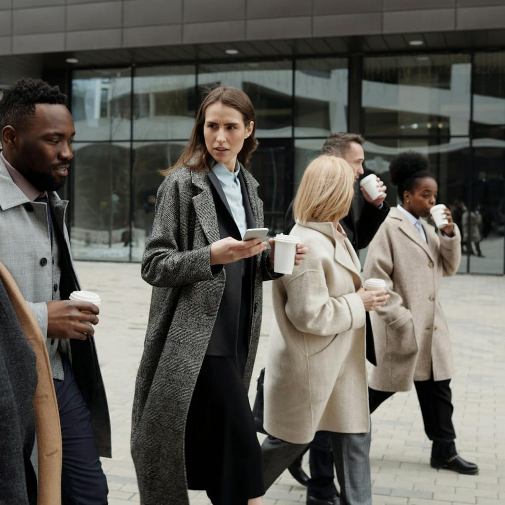 A group of people in smart coats holding take away coffee cups walk across a square outside. A large glass building is in the background.