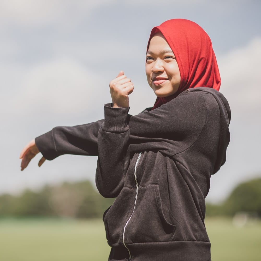 A woman in a red headscarf and black top stretching her arms, with some grey clouds in the background behind her.