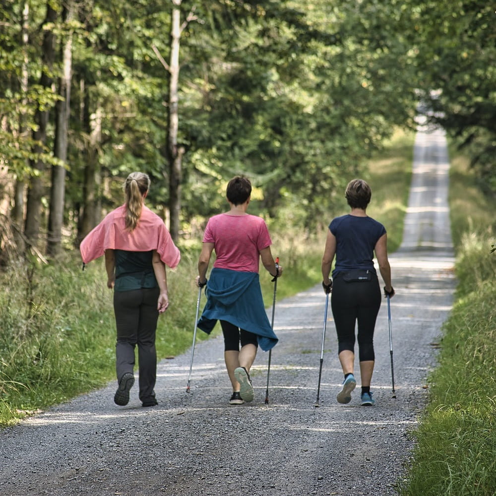 Three women walking in a horizontal line along a country road with grass and trees around them. Two of the women are using Nordic walking poles.