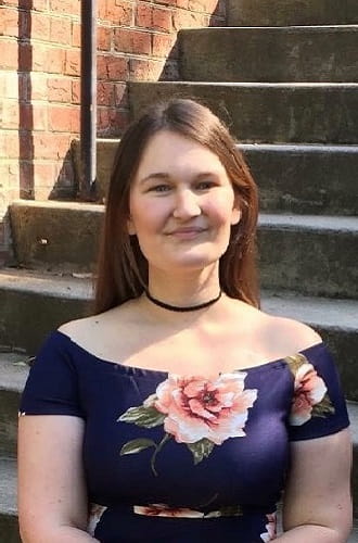 Johna, a white female with auburn hair, sits on a set of concrete stairs outdoors. She wears a blue dress with pink flowers. She is smiling.