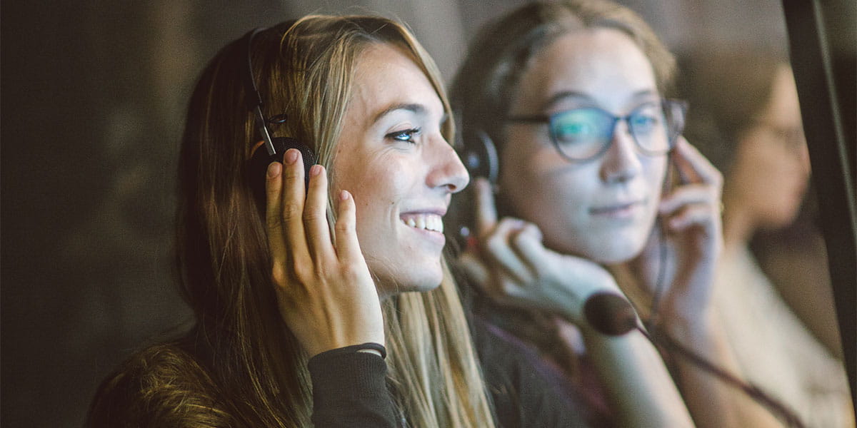 Two girls with headphones and microphone sets on looking at a computer screen.