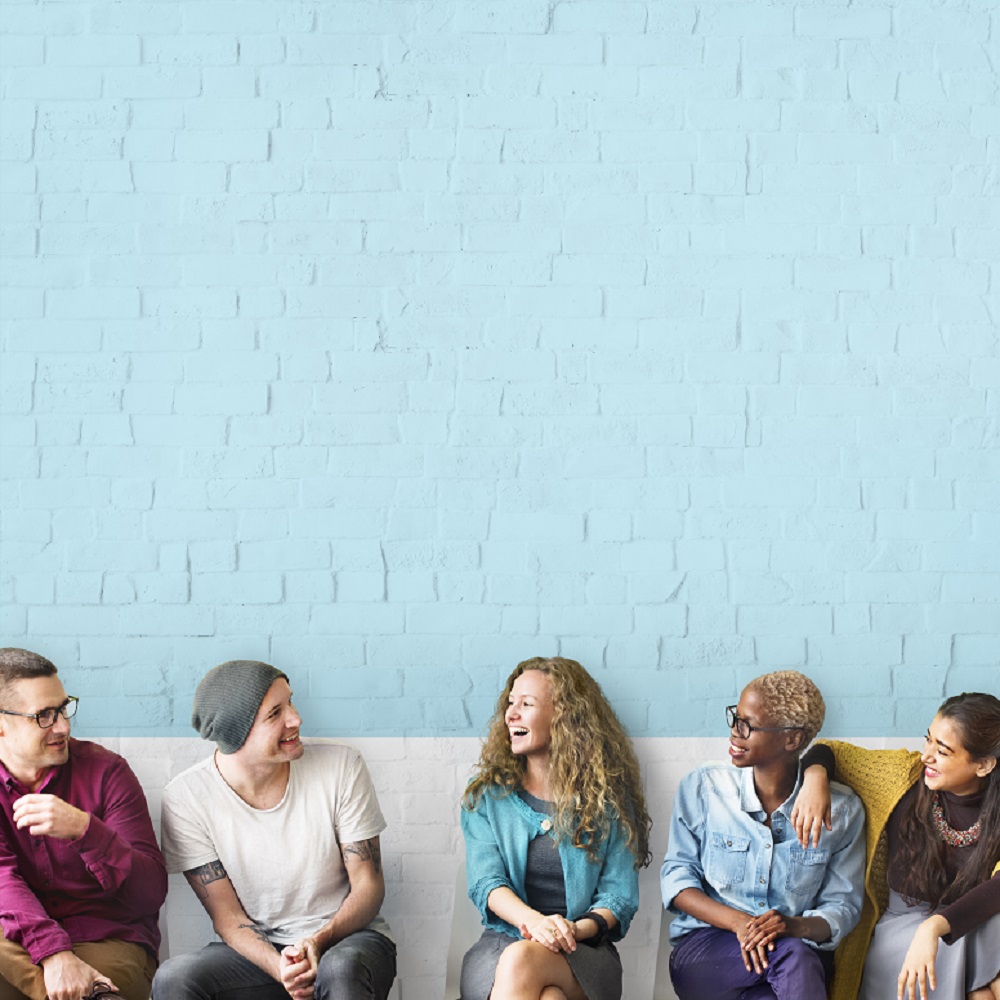 A group of friends sat in a line talking against a blue painted brick wall