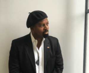 Ben is leant against a white wall. He is in a black suit and wearing a white shirt and black beret. 