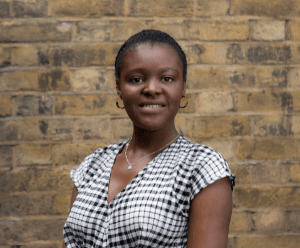 Chantelle is stood in front of  a brick wall. She is wearing a black and white checked top and is facing the camera, smiling.