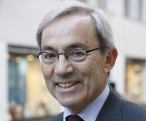 Christopher is wearing a grey suit with a burgundy tie, the setting is blurred behind him and he is smiling.