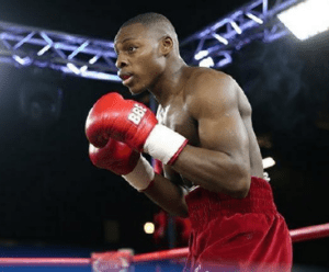 Dan is in a boxing ring, he is wearing red shorts and red boxing gloves. He is faced to the side, focussing ahead.