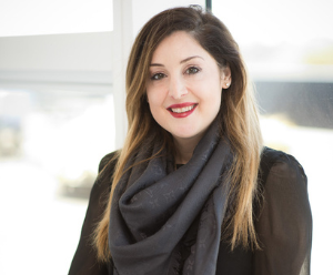 The image shows Elizabeth wearing a black dress and a grey scarf. She is stood in front of a window, the background is bright but blurred.