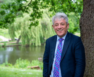 John is wearing a dark suit with a purple tie. He is stood next to a tree with green parkland behind him.