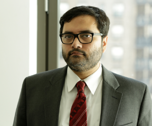 Monjurul is wearing a grey suit, white shirt and a red tie. The background is blurred.
