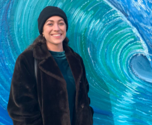 Rebecca is wearing a winter coat and a black beanie hat, she also has a bag over her shoulder. She is stood in front of a painting of a wave.