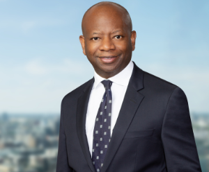 Trevor is wearing a navy suit and white shirt, and a navy tie. He is stood in front a blurred city backdrop.