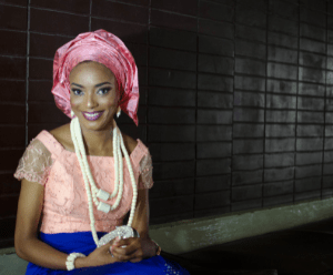 Ugonnaya is sat down, the background is black tiled wall. She is wearing a bright blue skirt, a peach top with chunky cream beaded necklace. She wears a rose pink head scarf and is smiling.