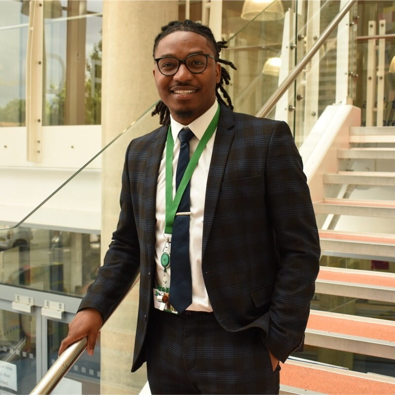 Johnoi stands on a flight of stairs wearing a suit and tie, a green lanyard is around his neck.