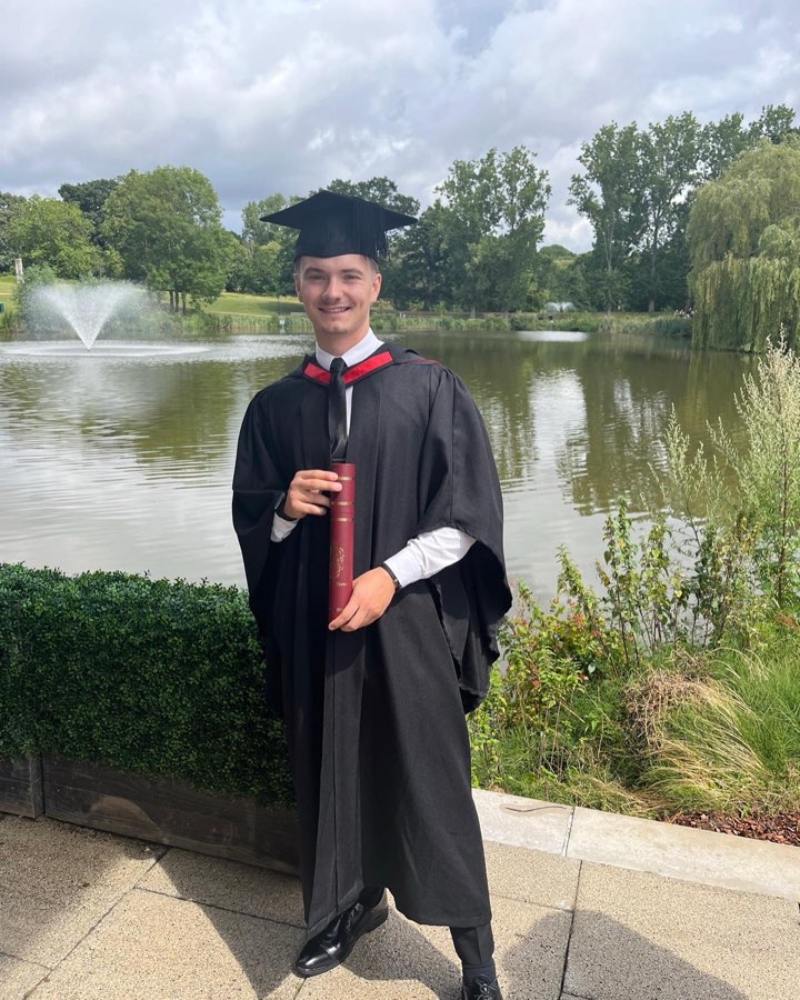 Student George wearing a graduation gown and holding his diploma in front of a lake