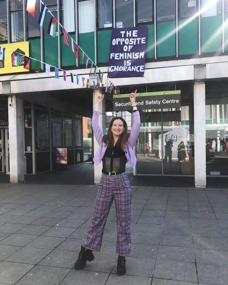 Marina Cusi Sanchez stands on Square 3 outside the Security Helpdesk pointing to a banner which reads "The opposite of feminism is ignorance".