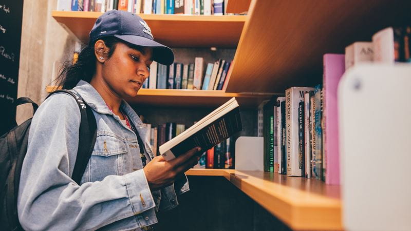 A young south Asian woman wearing a blue baseball cap reads a book in the library