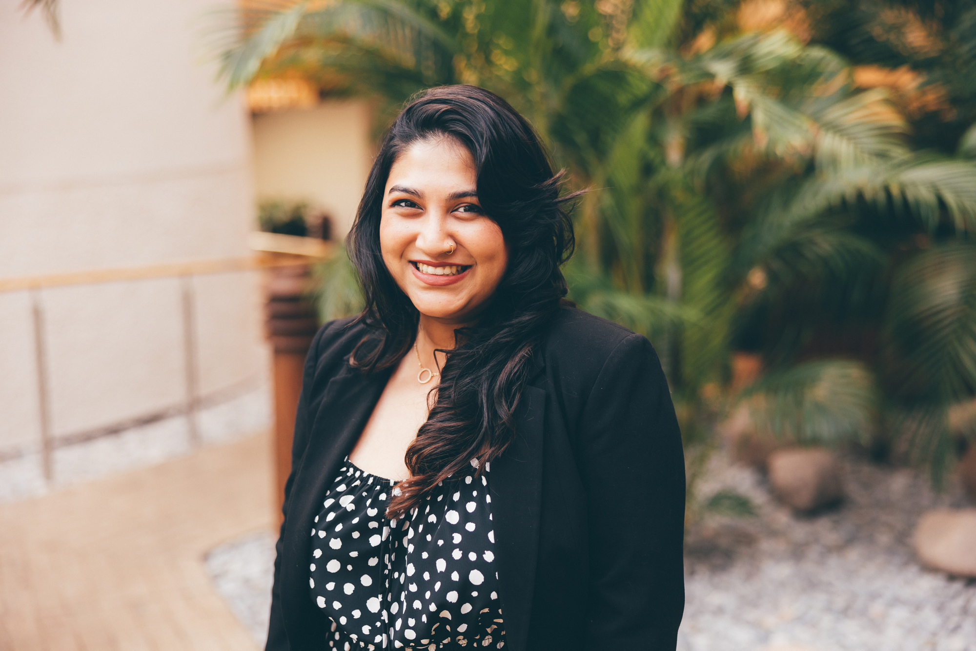 Sharina Islam, MBA 2022 graduate, stands in the Essex Business School building indoor winter garden. She wears a black suit jacket over a black and white spotted blouse and wears her long black hair loose over her shoulders.