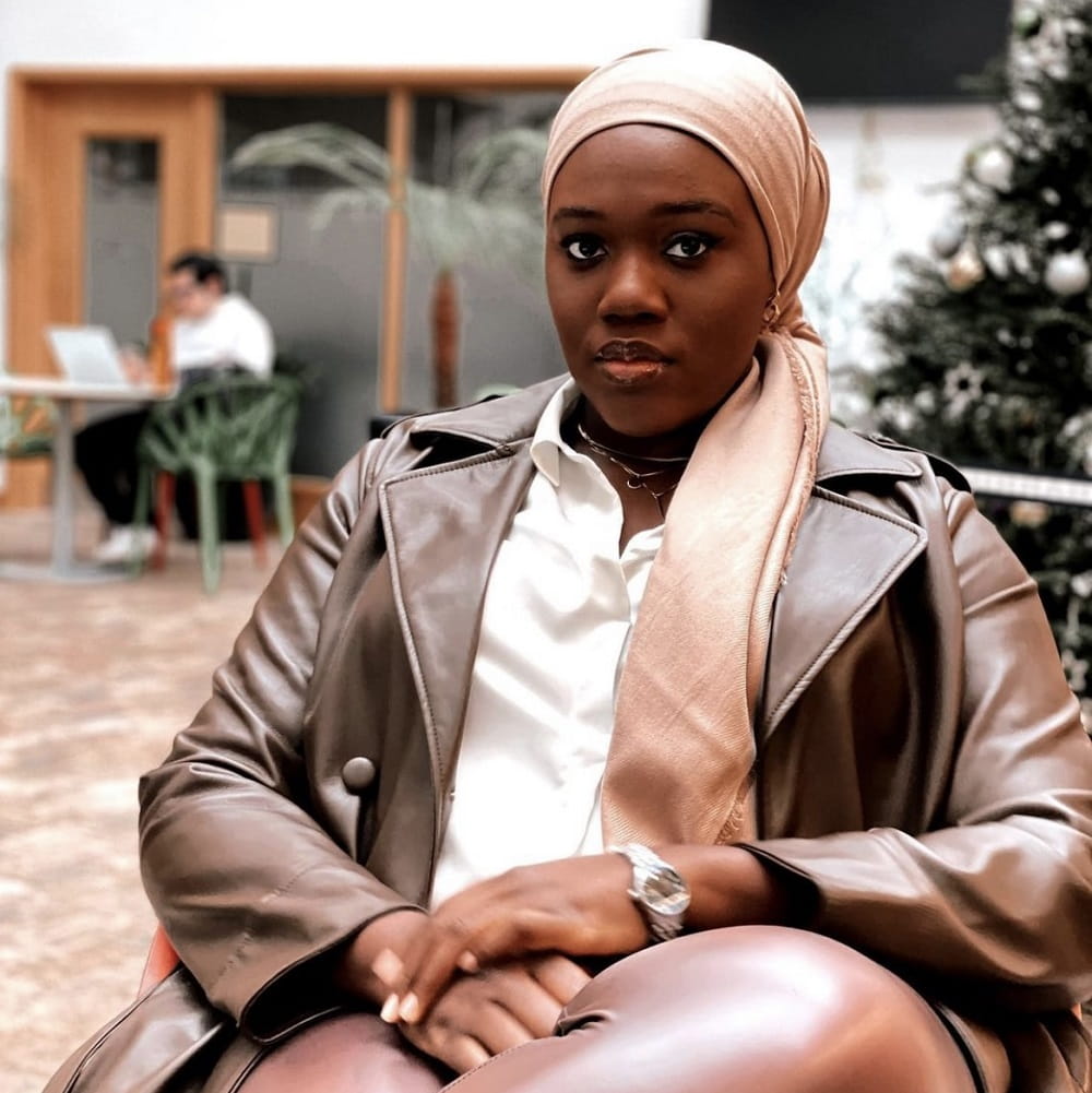 Habibat Kawu, a Black woman wearing a headscarf and brown leather jacket, sitting indoors. There is a person on a laptop in the background behind her shoulder.