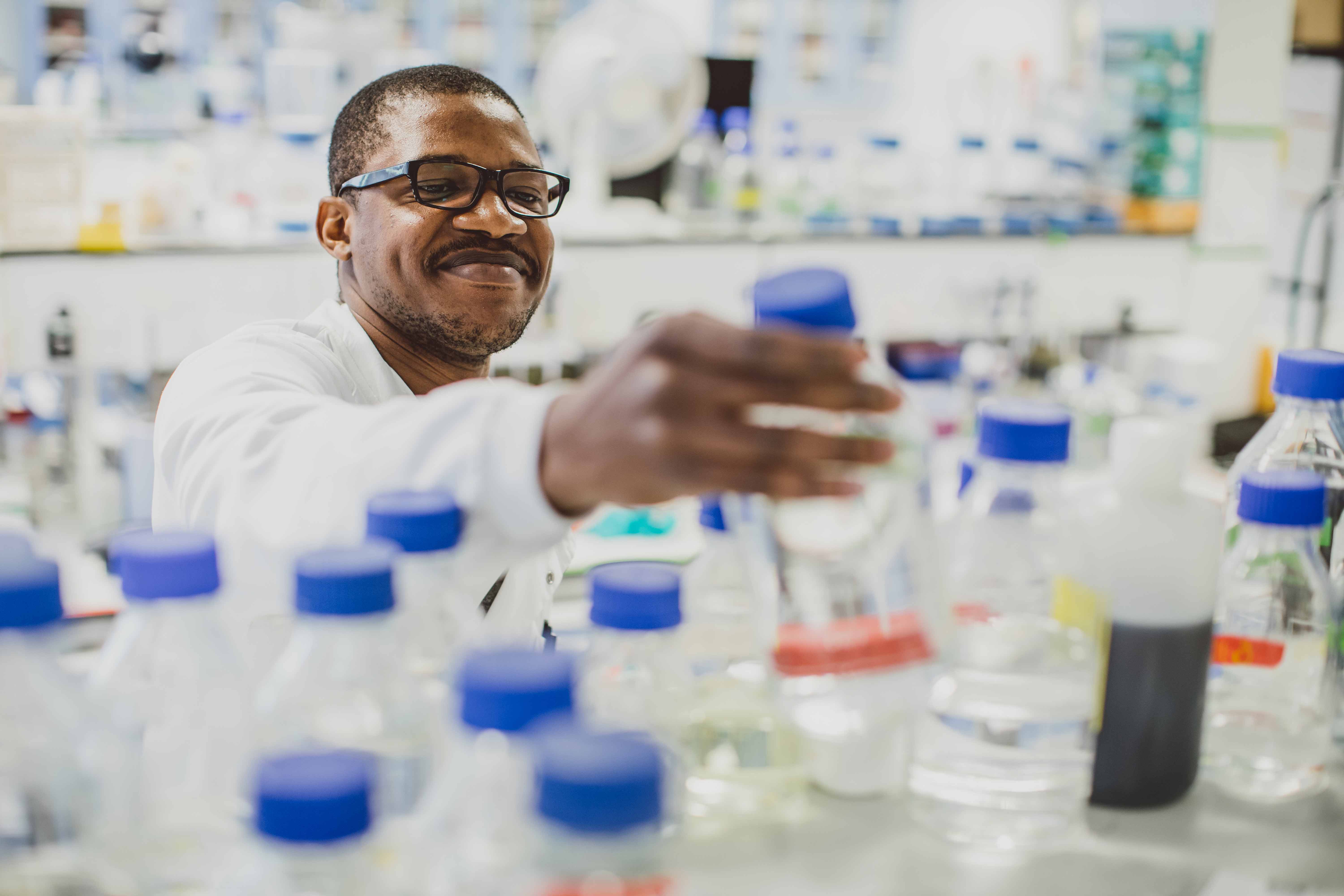 A male scientist in a lab, reaching for a bottle in the foreground