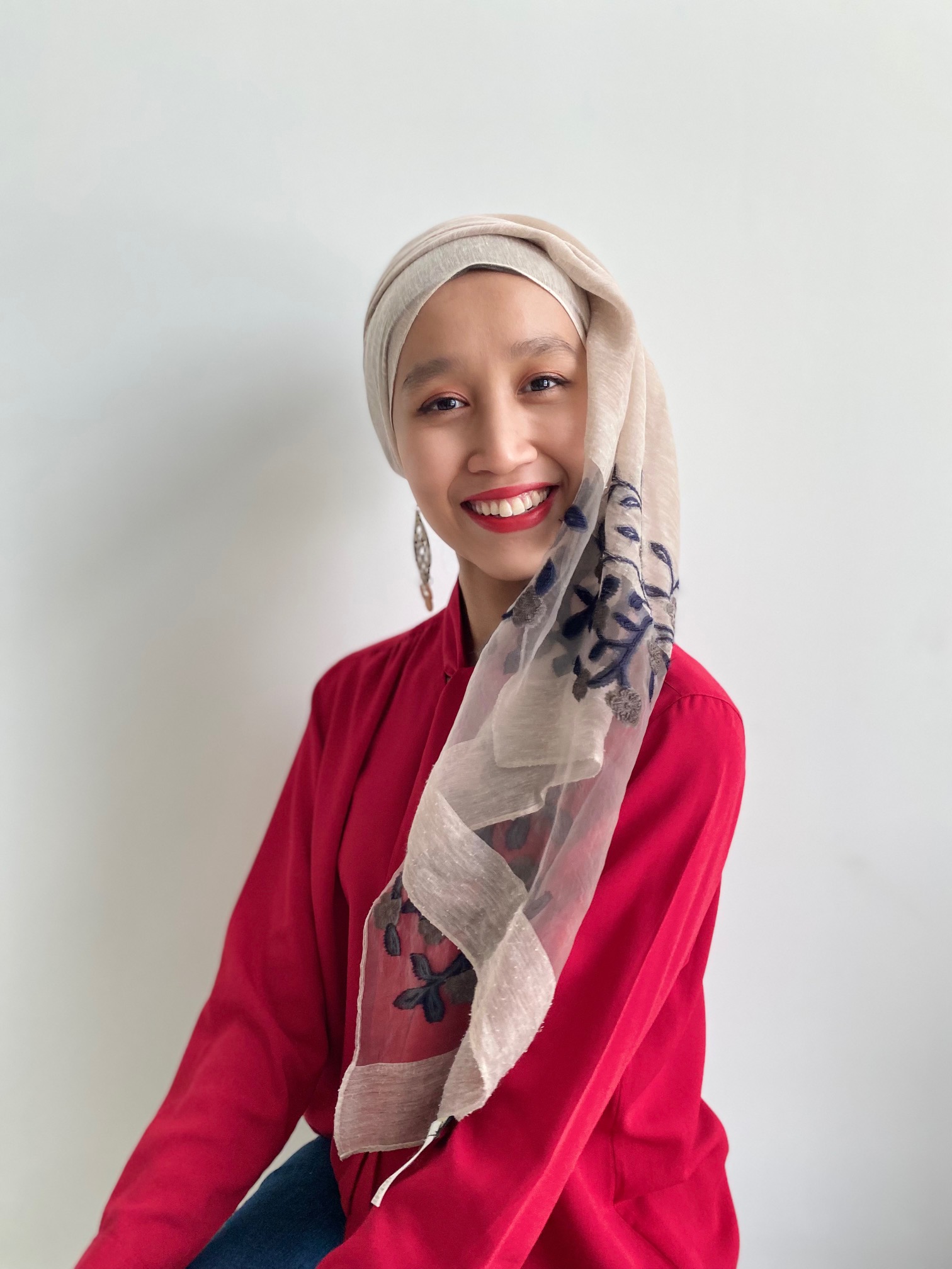 Raudhah facing the camera and smiling, she is sat in front of a white wall.