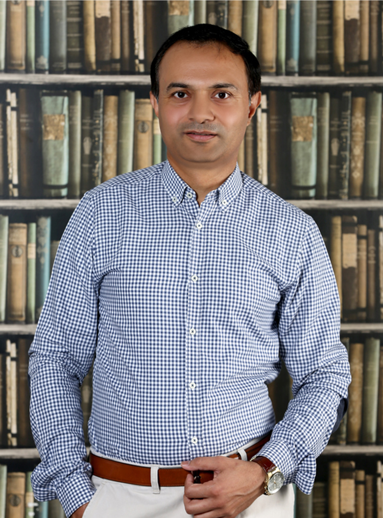 Mandeep Sandhu, MBA 2015 Alumni and CEO and Founder of AirLuxxis Aviation Services, stands wearing a shirt and chinos with a brown belt in front of a bookcase filled with old books.