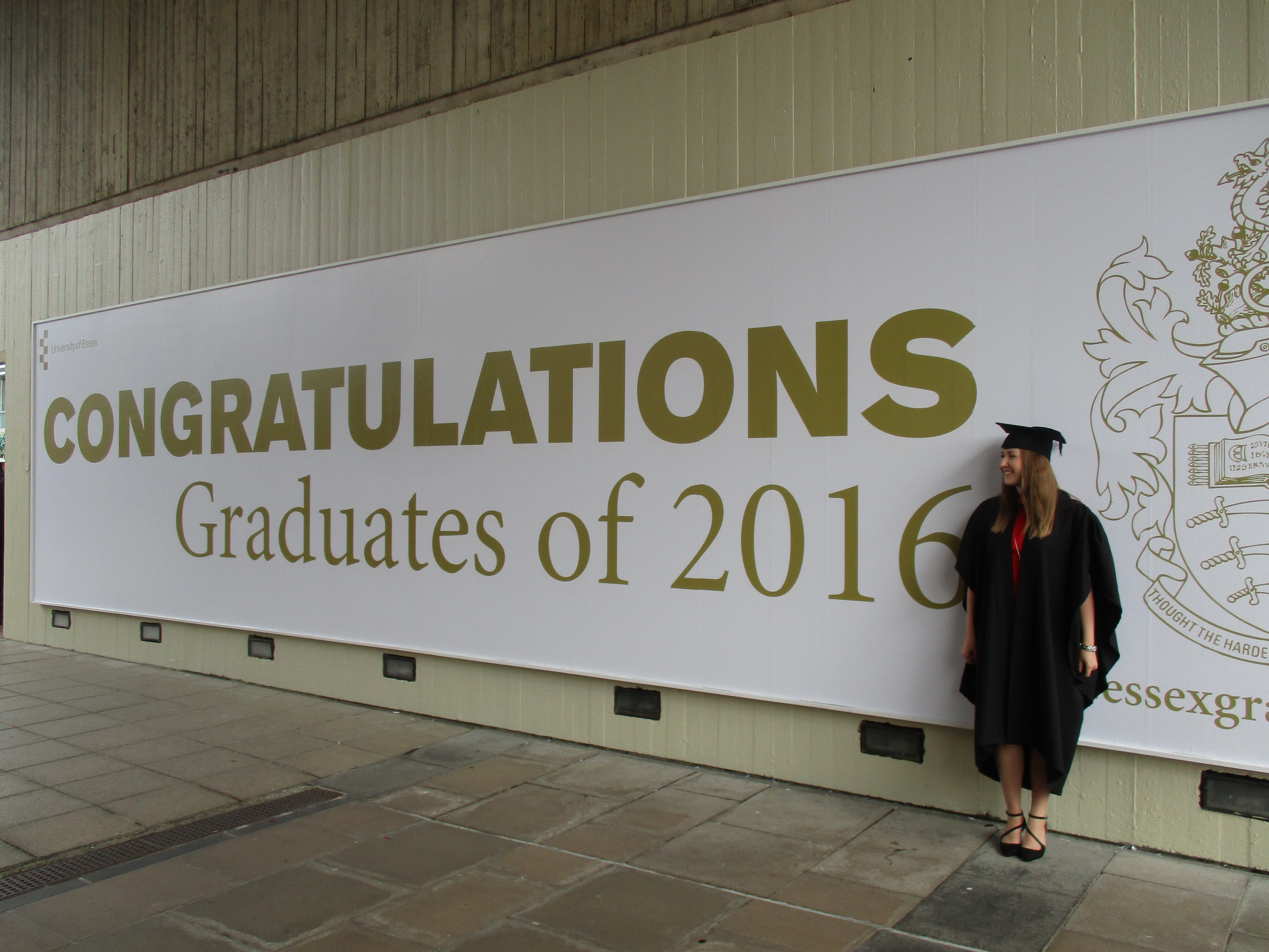 Alice on her graduation day, dressed in robes and stood in front of a graduation poster on our Colchester Campus