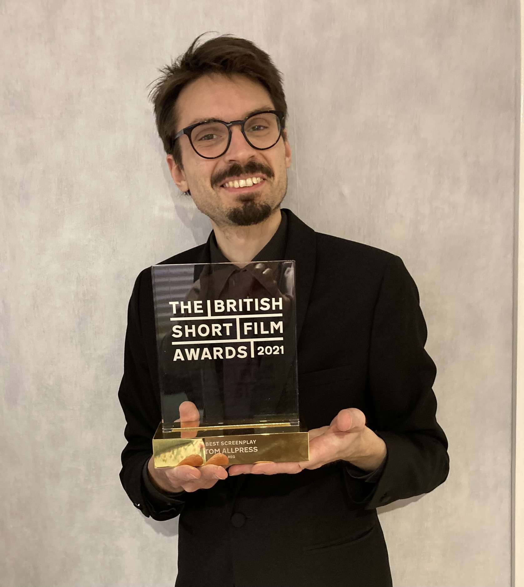 Thomas smiling, holding his Best Screenplay award from the British Short Film Awards. The backdrop is a textured wall.