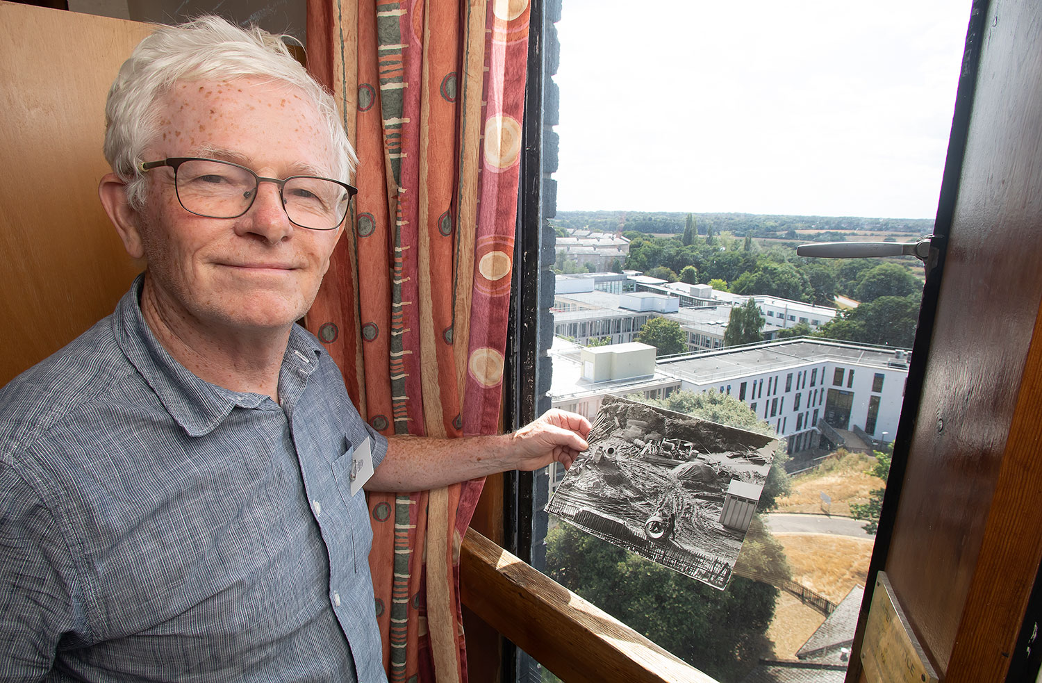 An alumnus stands by a window in Rayleigh Tower holding a black and white photo of the view that used to be outside.